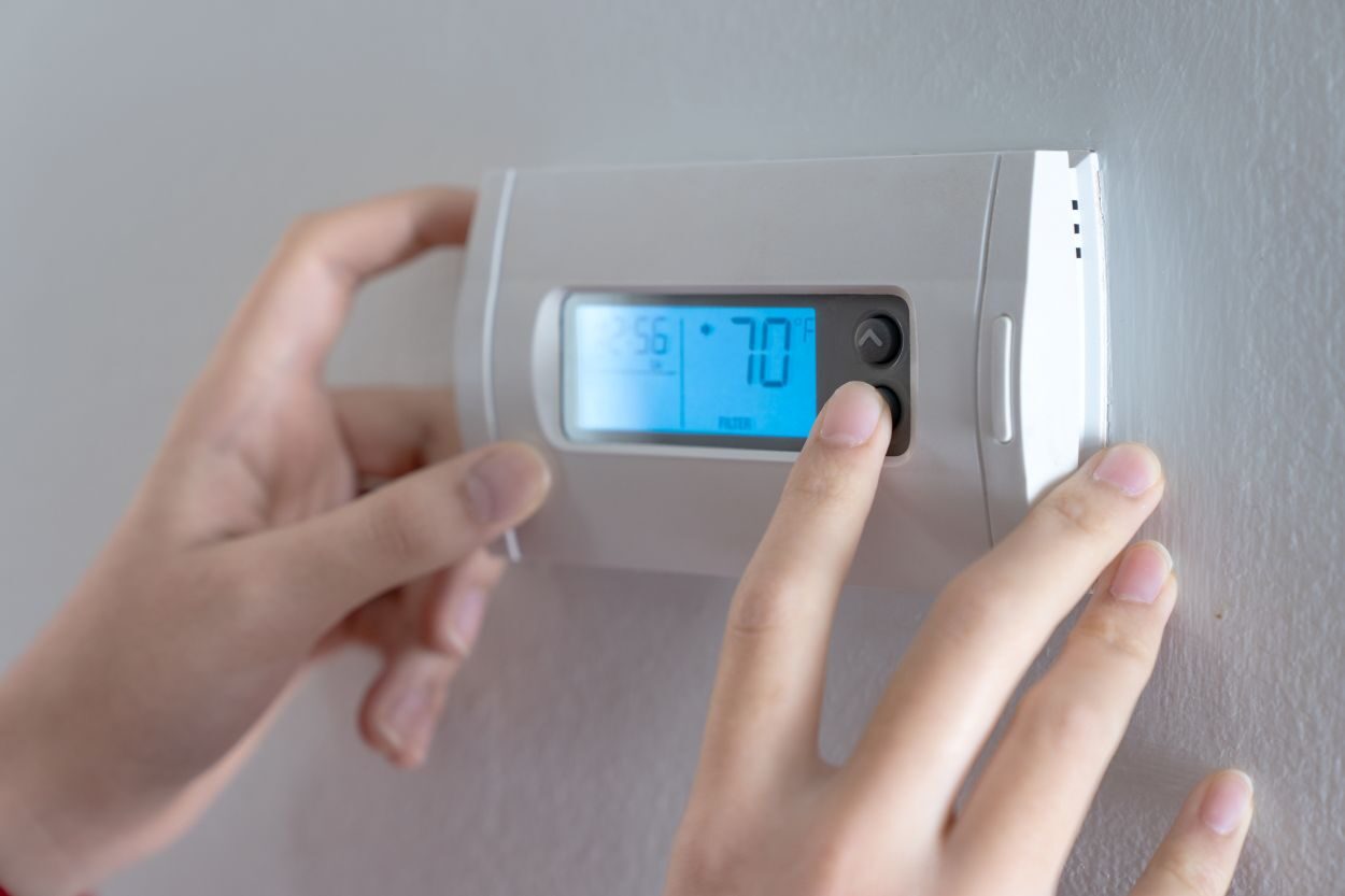 A woman is pressing the down button of a wall attached house thermostat with digital display showing temperature 70 degree Fahrenheit for heating, cooling, electricity and gas saving See Less
